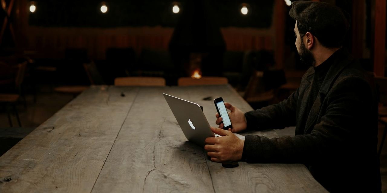 Someone answering messages on a phone and laptop at a long table in a quiet restaurant at night